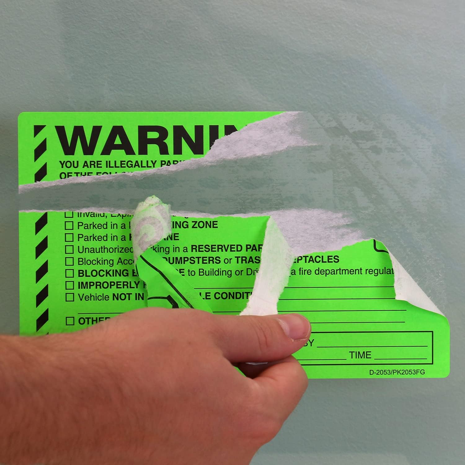 A hand peeling off a partially torn green parking violation sticker from a car window.