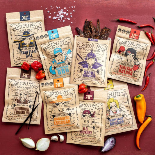 Eight bags of Righteous Felon beef jerky arranged on a dark red surface with food props including tomatoes, garlic, onions, peppers, and vanilla beans. The bags feature different illustrated character designs and flavor names such as Truffule Bill, Seoul Survivor, and Bourbon Frankin.