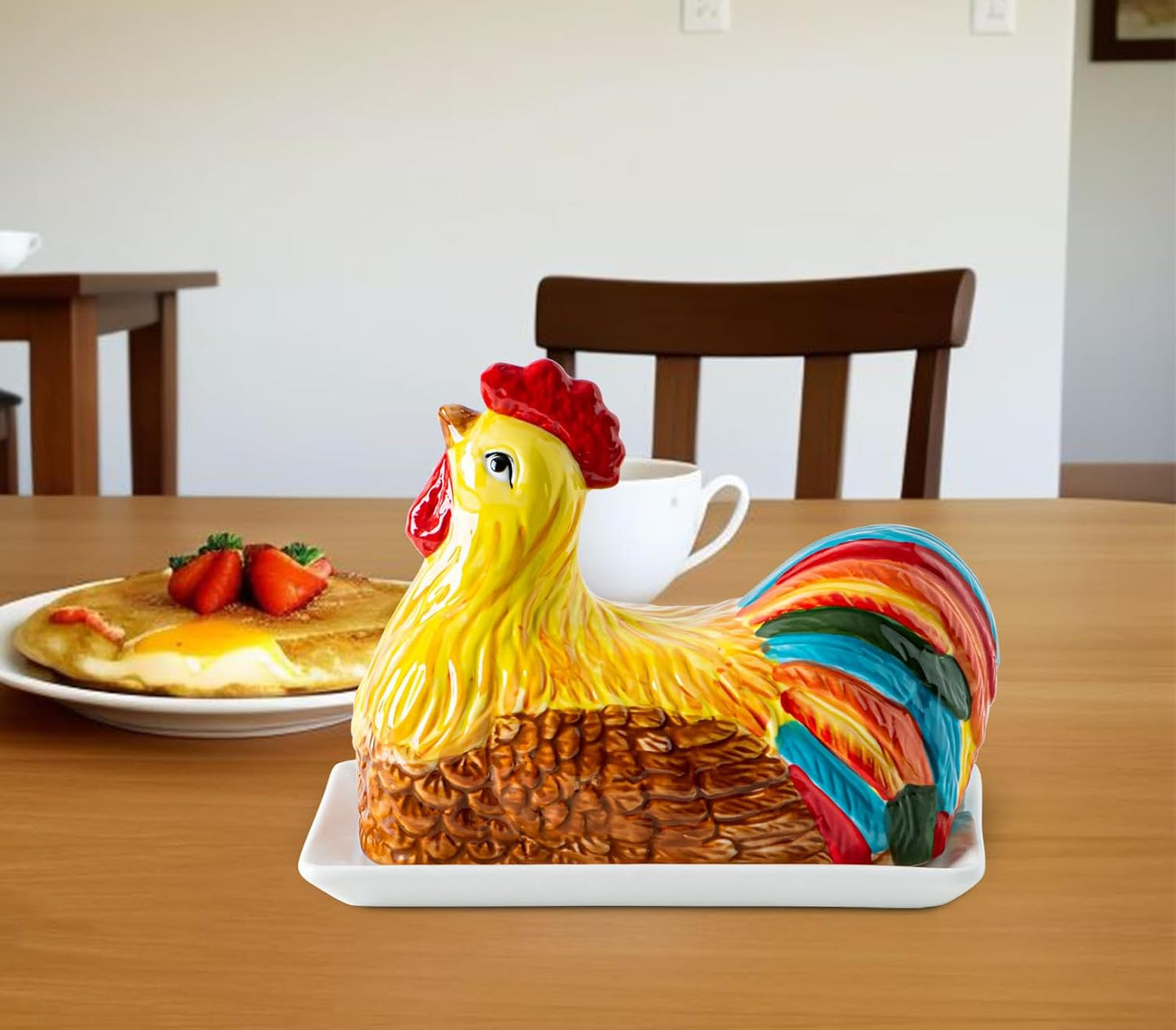 Colorful ceramic chicken-shaped butter dish on a white tray, on a wooden table with a plate of pancakes, strawberries, and a white teacup in the background.