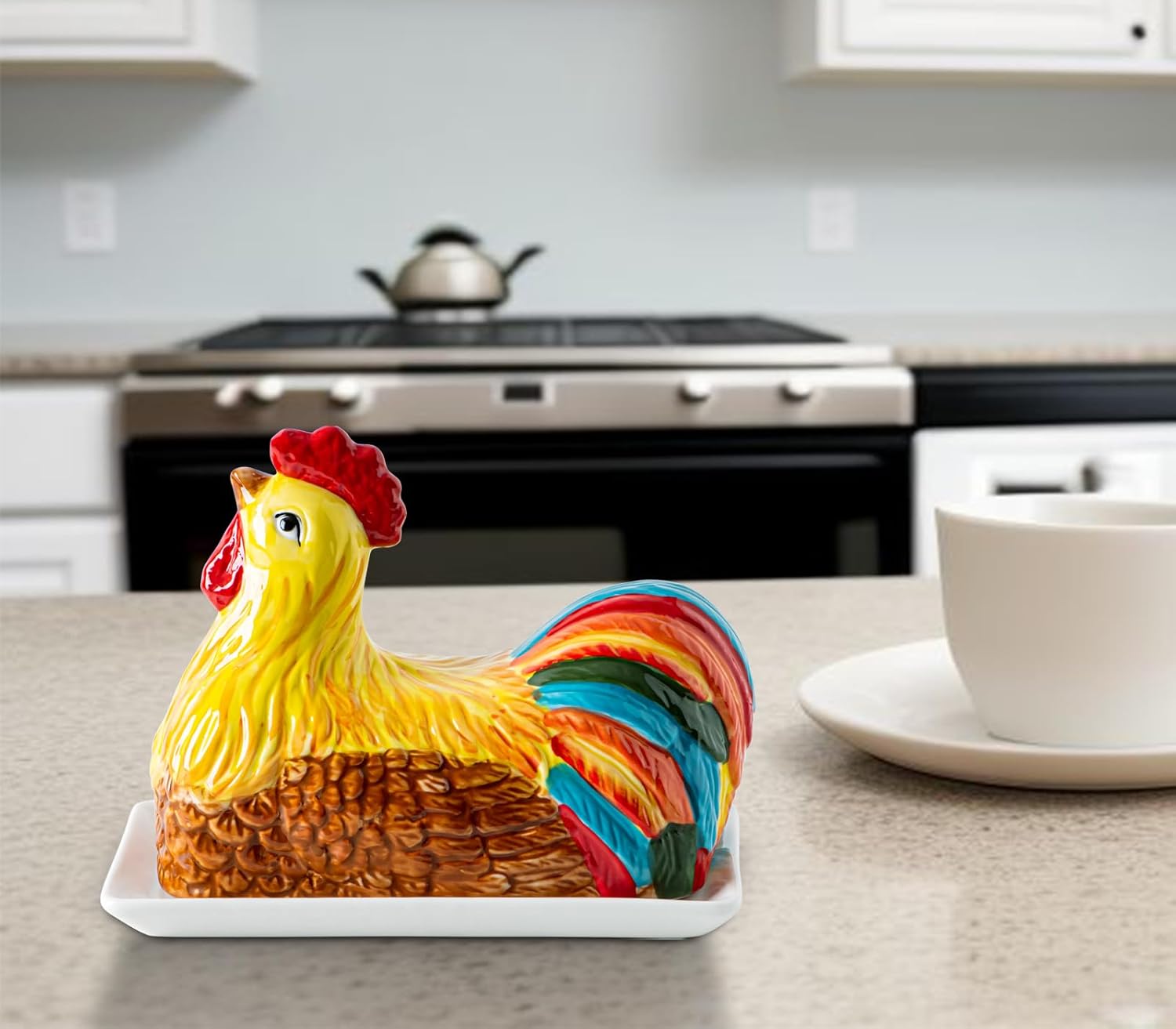 Colorful ceramic chicken-shaped butter dish on a white tray, on a kitchen counter with a stove, kettle, and white cup on a saucer in the background.