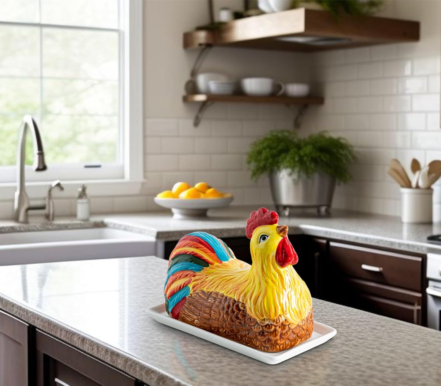 Colorful ceramic chicken-shaped butter dish on a white tray in a modern kitchen with a sink, citrus bowl, potted plant, and wooden shelves in the background.