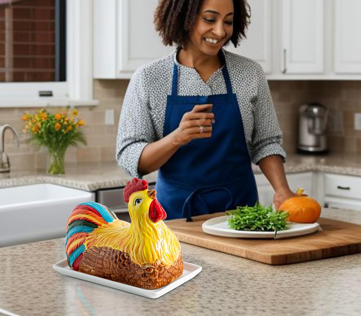 A person in a blue apron stands in a kitchen with a colorful ceramic chicken-shaped butter dish on a white tray, a plate of greens, a pumpkin, and flowers in the background.
