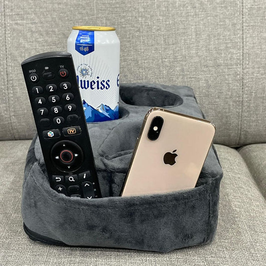 A gray plush TV pillow couch caddy organizer sitting on a light gray fabric couch, holding a black remote control, a can of Weiss beer, and a gold iPhone.