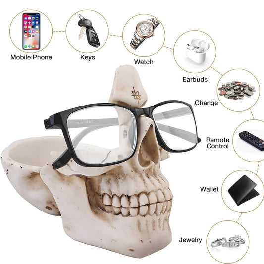 A skull-shaped eyeglasses holder displaying black-framed glasses, with circular callouts around it showing items that can be stored, including a mobile phone, keys, watch, earbuds, cash, remote control, wallet, and jewelry.