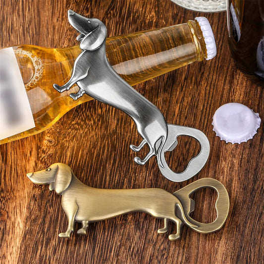 Two dachshund-shaped bottle openers (silver and bronze) on a wooden surface with a beer bottle, a bottle cap, and a dark bottle in the background.