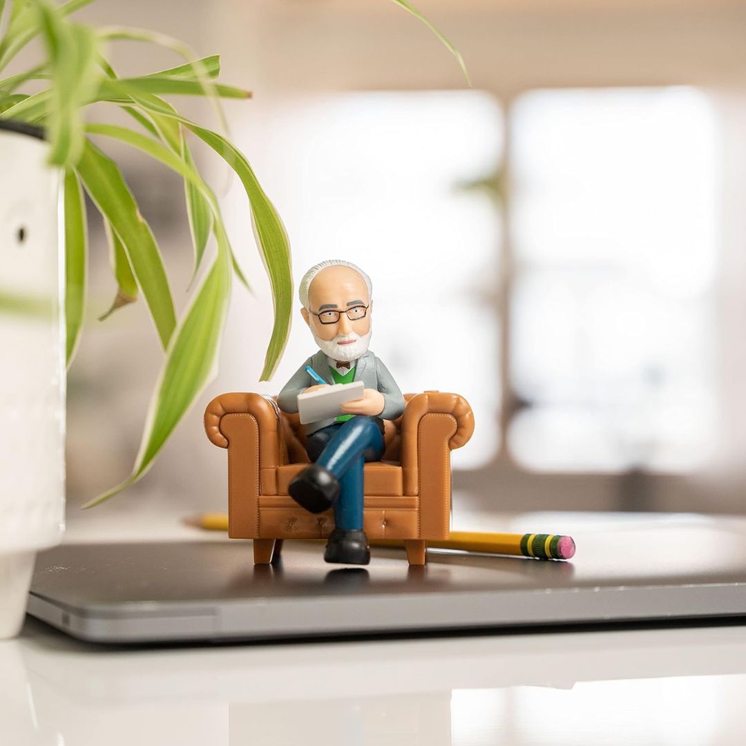 Figurine of a male therapist sitting on a couch with a book, surrounded by a plant and a pencil on a desk.