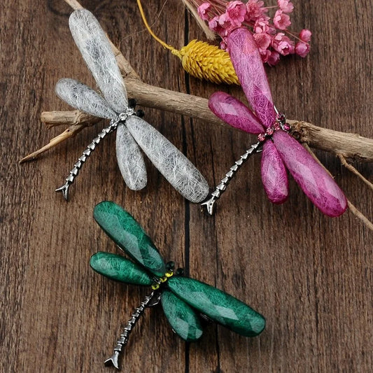 Three dragonfly brooches in silver, pink, and green with gemstone wings on a wooden surface, with dried flowers in the background.