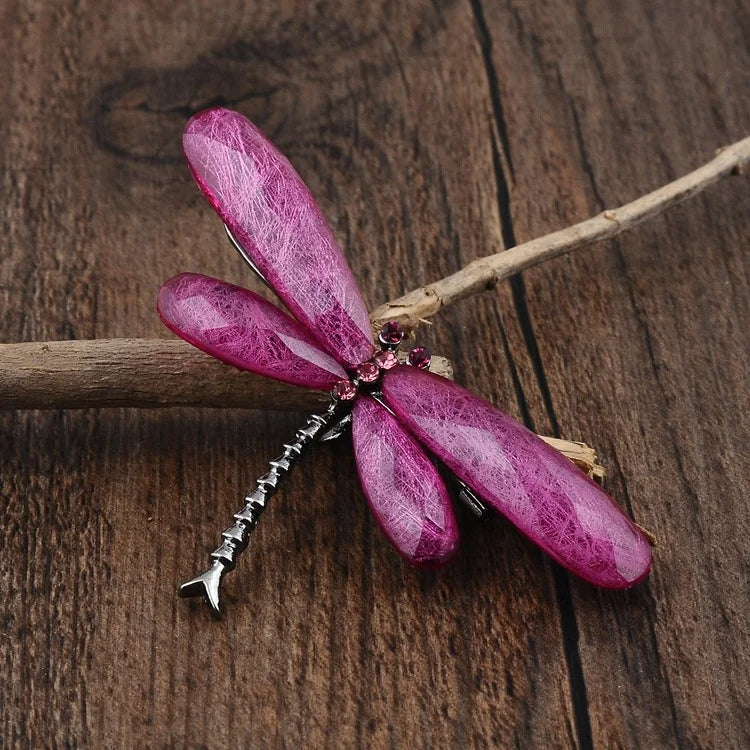 A pink dragonfly brooch with gemstone wings, adorned with pink gems, resting on a wooden branch against a wooden surface.