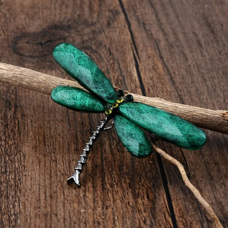 A green dragonfly brooch with gemstone wings, decorated with yellow and black gems, perched on a wooden branch against a wooden surface.
