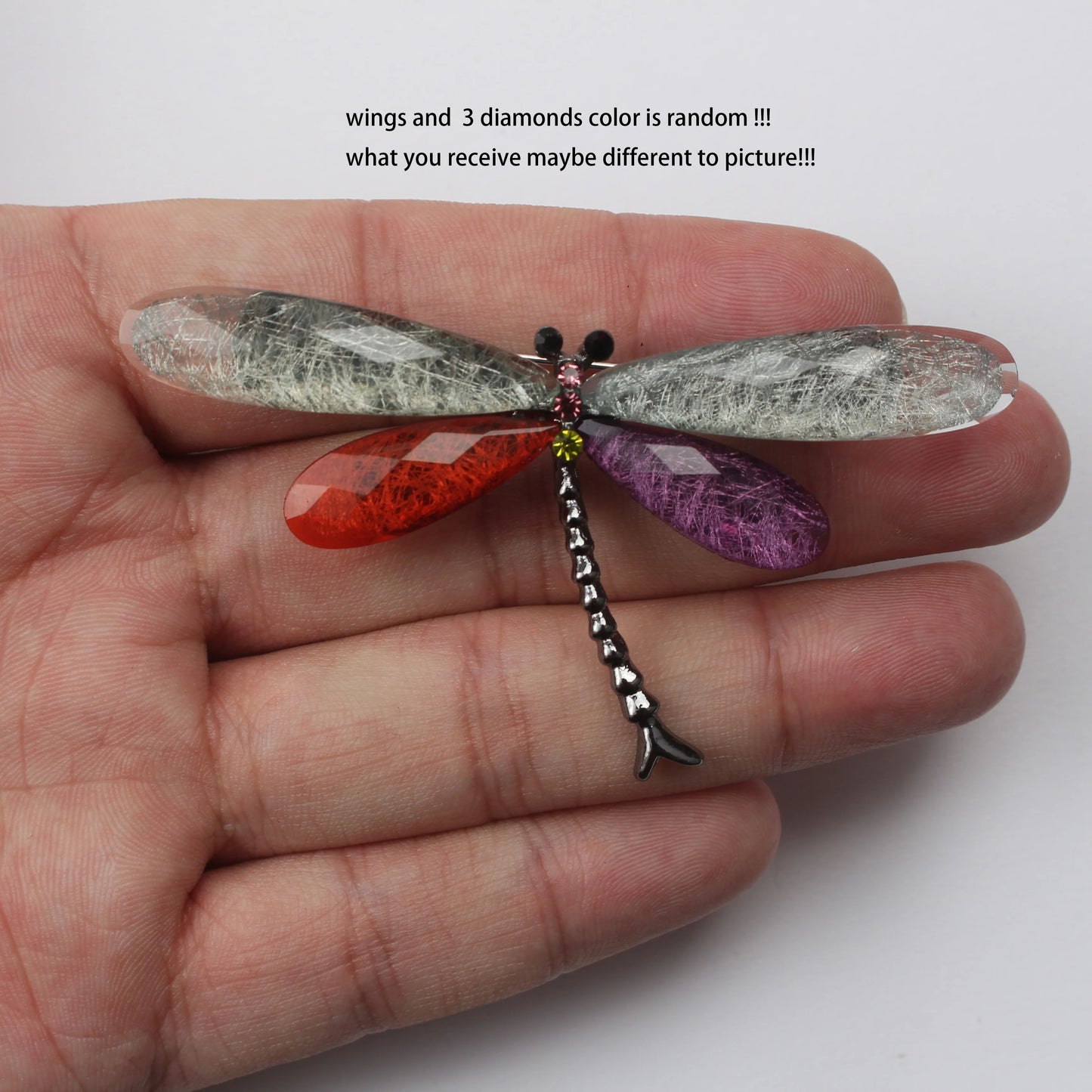 A dragonfly brooch with gray, red, and purple gemstone wings, held in a hand. Text above reads: "wings and 3 diamonds color is random!!! what you receive may be different to picture!!!"