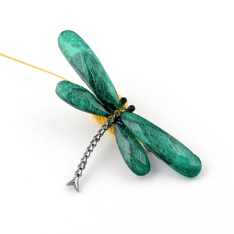 A green dragonfly brooch with gemstone wings, decorated with yellow and black gems, on a white surface with dried yellow foliage in the background.