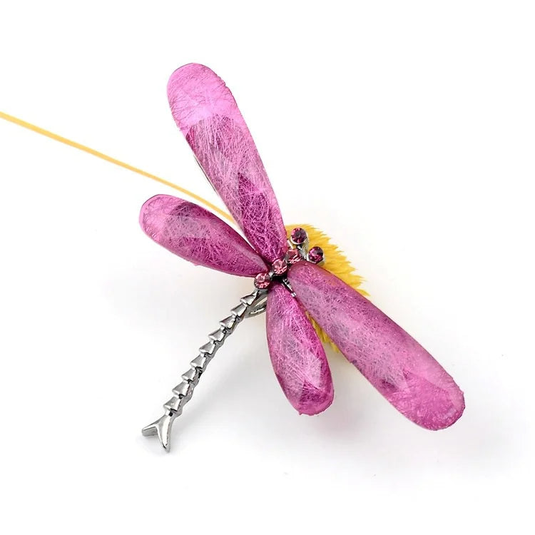 A pink dragonfly brooch with gemstone wings, adorned with pink gems, resting on a wooden branch against a wooden surface.