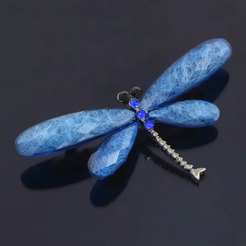 A blue dragonfly brooch with gemstone wings, adorned with blue and black gems, against a dark reflective surface.
