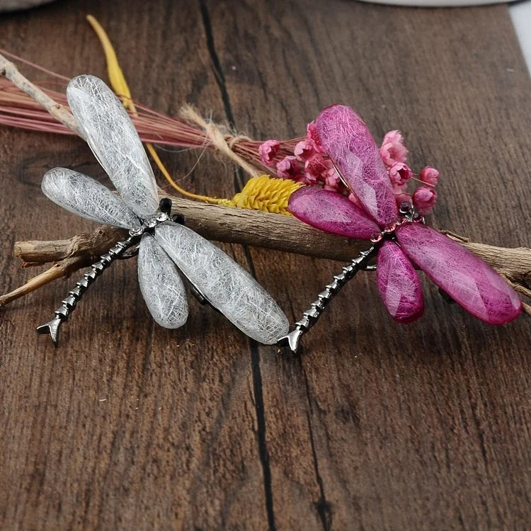 Two dragonfly brooches, one gray and one pink with gemstone wings, adorned with gems, resting on a wooden branch with dried flowers on a wooden surface.