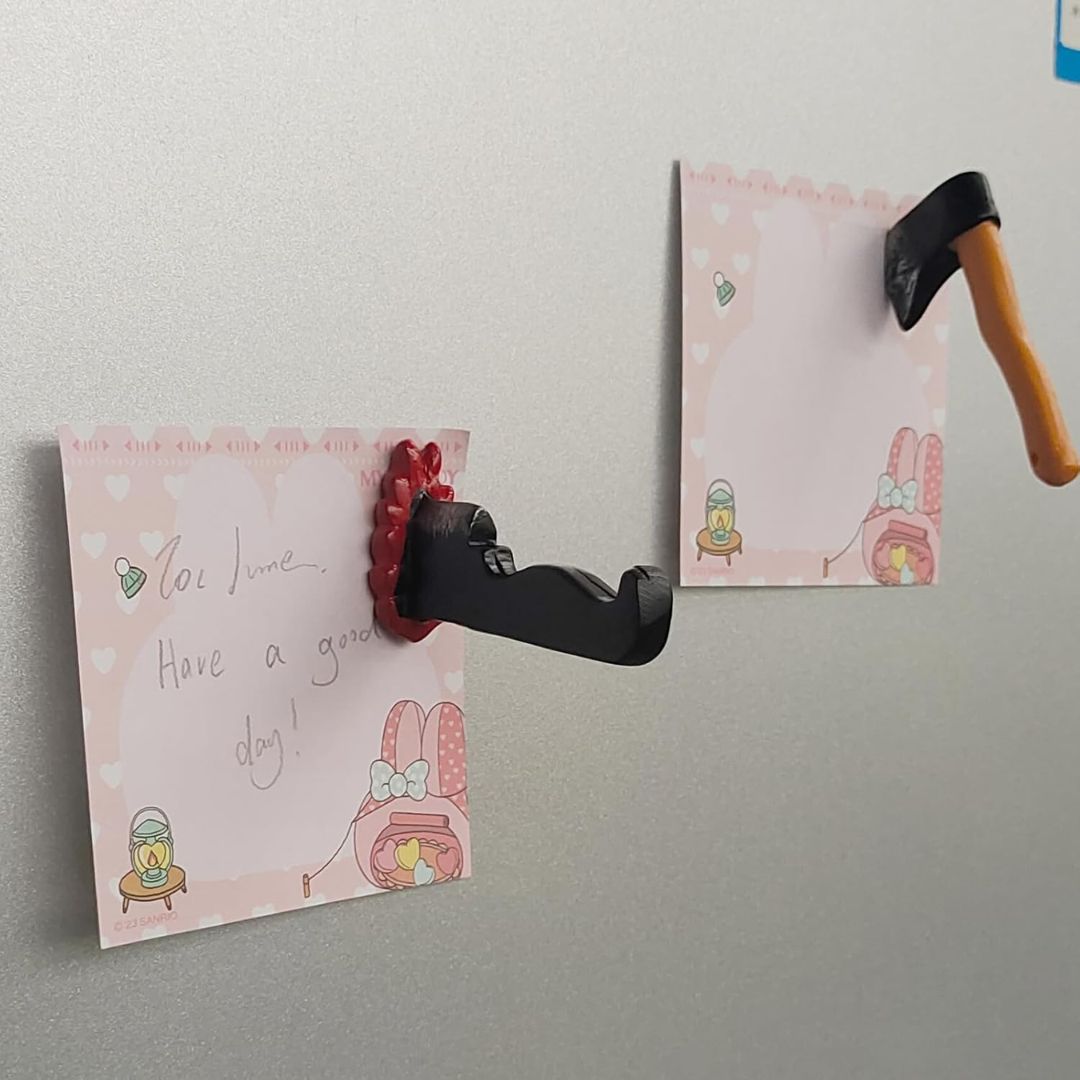 Two funny fridge magnets shaped like a small axe and a bloodied knife, shown attached to pink heart-patterned note cards.