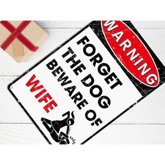 Funny metal warning sign reading “Forget the dog beware of wife” displayed on a wooden surface with a gift box
