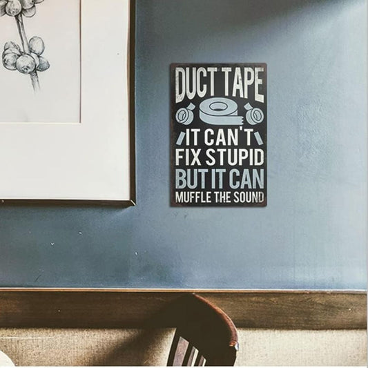A rectangular wall sign with the text 'DUCT TAPE IT CAN'T FIX STUPID BUT IT CAN MUFFLE THE SOUND' in white lettering on a dark charcoal background, mounted on a blue-gray wall in a room setting.