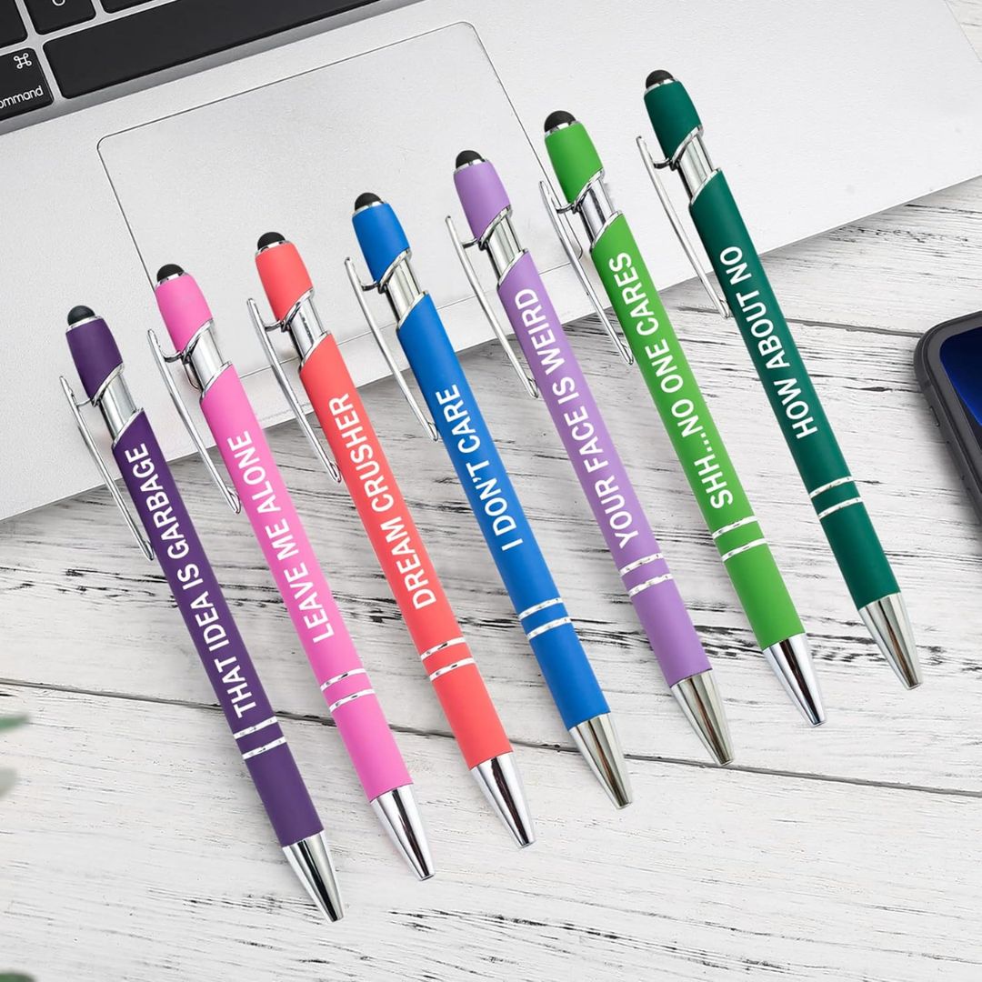 A set of colorful pens with sarcastic text on a desk with laptop,.