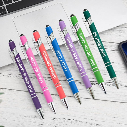 A set of colorful pens with sarcastic text on a desk with laptop,.