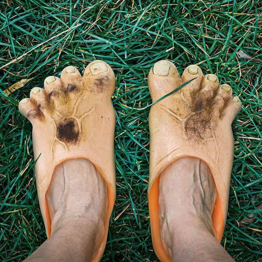 A person's feet wearing a pair of slippers that look like hobbit feet standing on grass