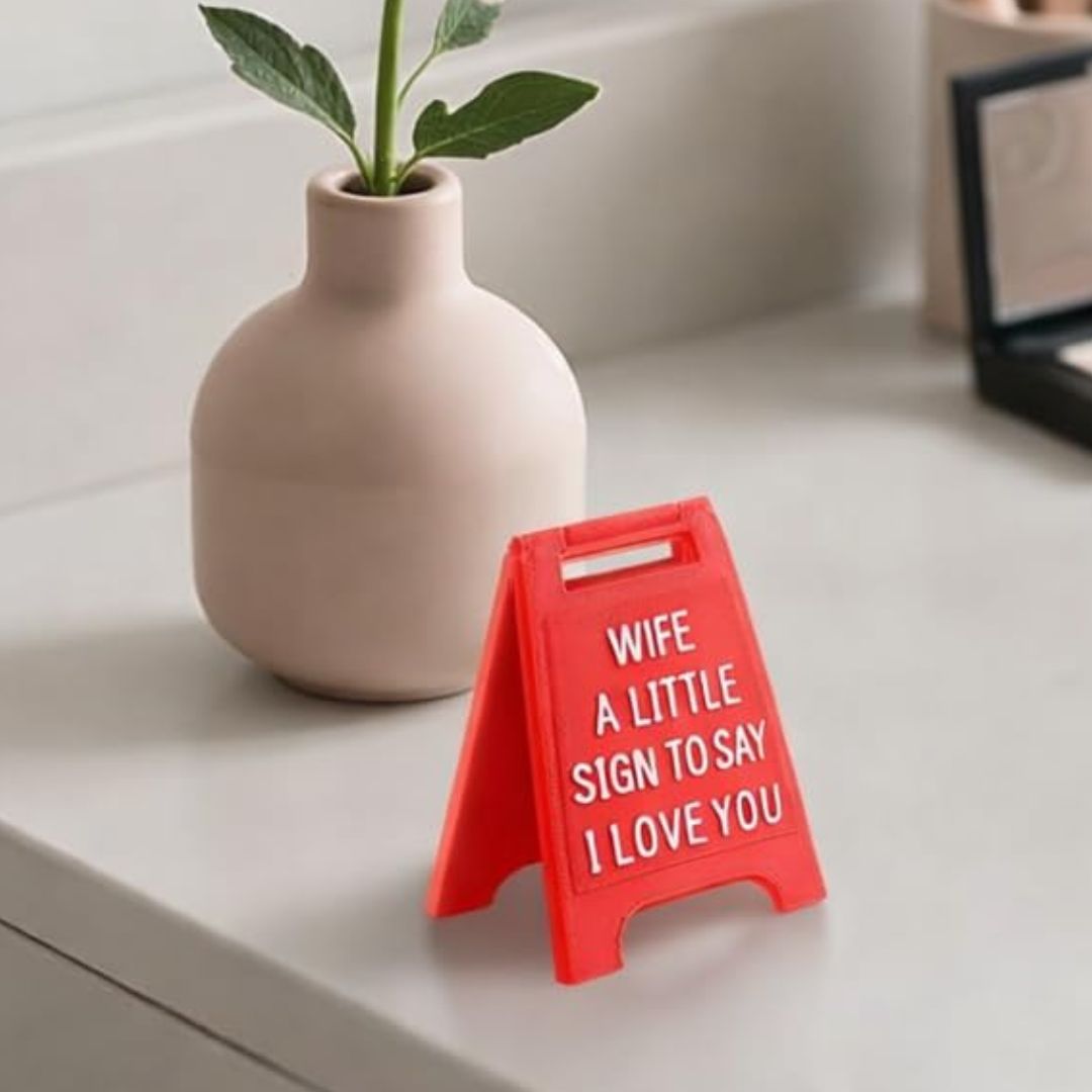 A small red A-frame sign with white text reading 'WIFE A LITTLE SIGN TO SAY I LOVE YOU' sits on a light countertop beside a matte beige vase with a green plant.