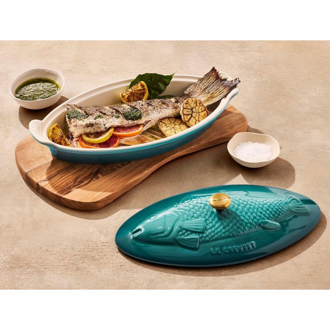 Le Creuset Riviera stoneware fish baker with roasted whole fish, citrus and garlic, with fish-shaped lid beside it