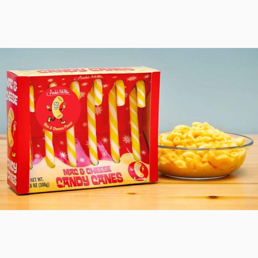 Box of Mac & Cheese Candy Canes next to a bowl of macaroni and cheese on a wooden surface.