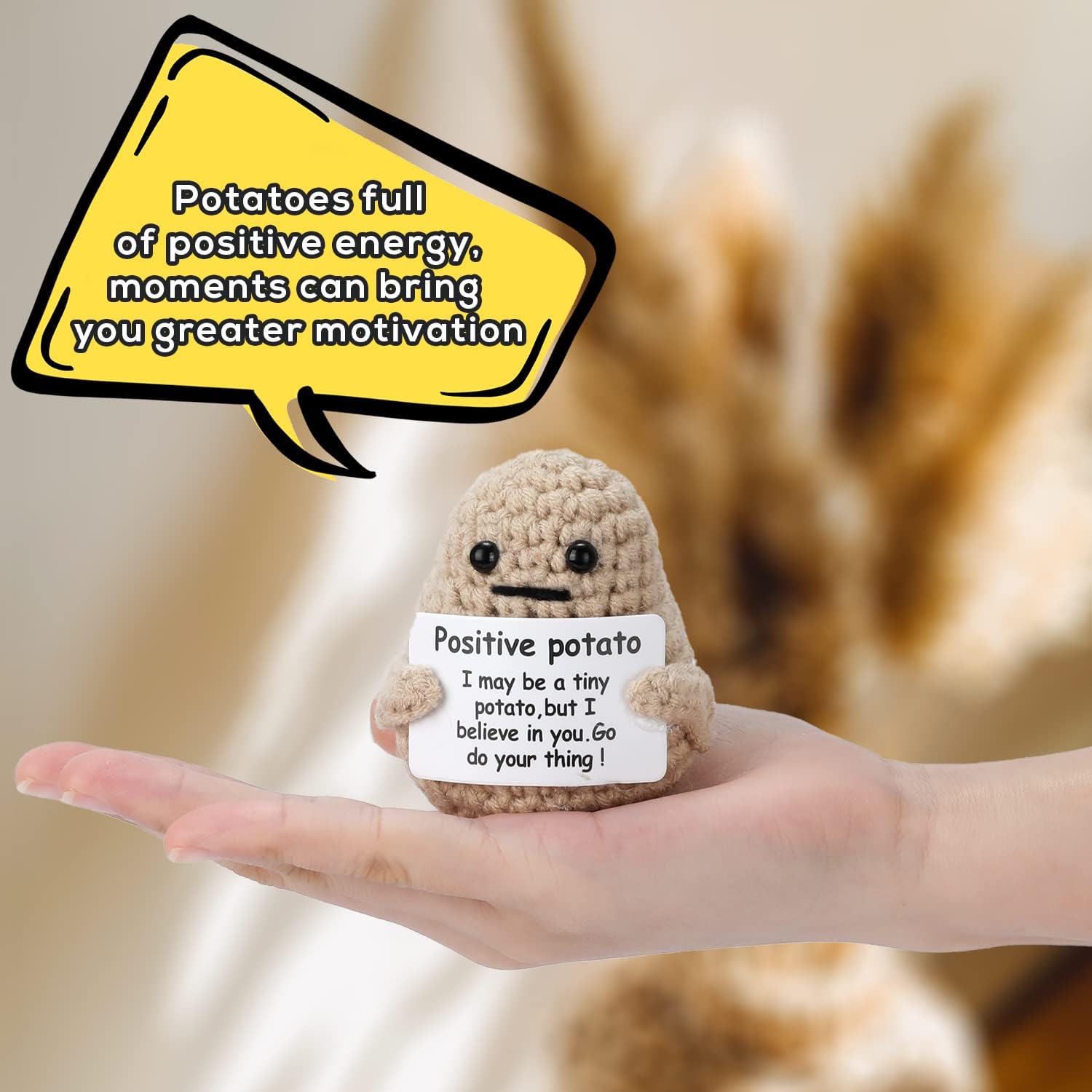 A small, crocheted plush toy shaped like a potato with black button eyes and a simple stitched mouth is sitting on an open human hand. The plush toy holds a white card with a motivational message that reads: "Positive potato. I may be a tiny potato, but I believe in you. Go do your thing!" A large yellow speech bubble above the toy contains another motivational message: "Potatoes full of positive energy, moments can bring you greater motivation." The background is blurred with warm tones, creating a cozy an