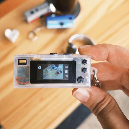 A hand holding a small retro digital camera, shown against a blurred wooden table background.