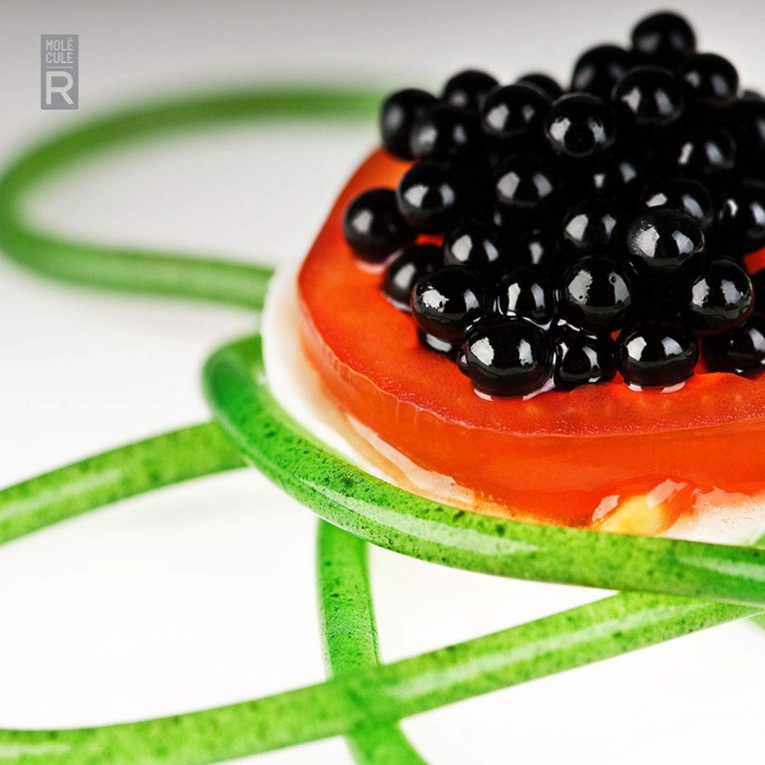 Molecular gastronomy dish featuring glossy black spheres on a red gel base with decorative green jelly strands