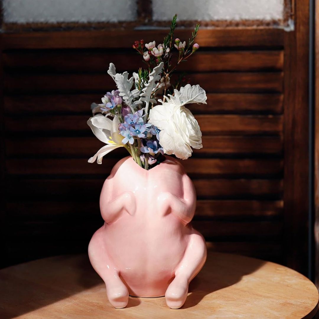 A glossy pink ceramic raw chicken shaped vase filled with white, blue, and purple flowers, set on a wooden table against a dark wooden slatted background.