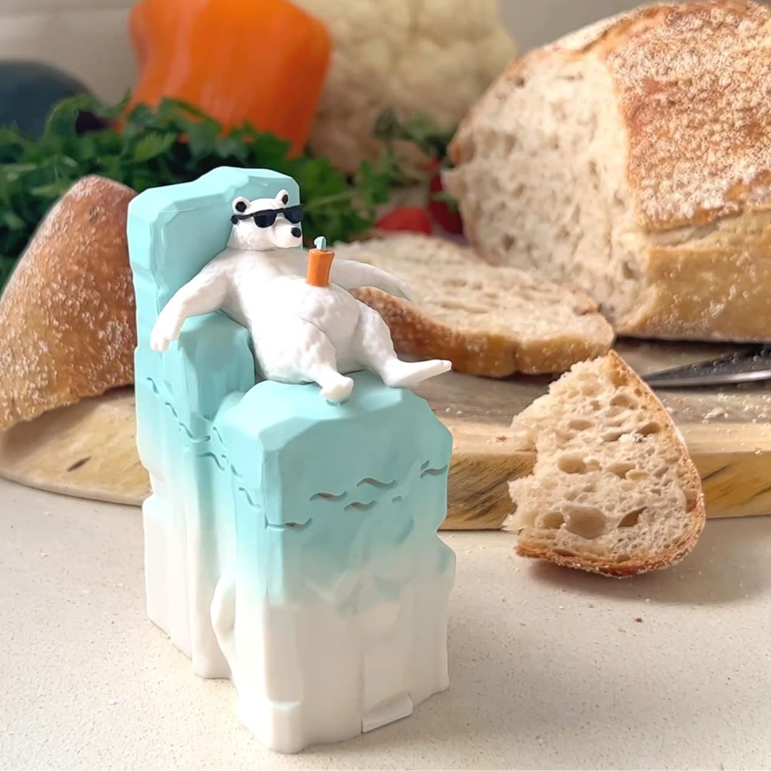 A small white polar bear fridge deodorizer wearing black sunglasses and holding an orange drink, reclining on a light blue and white iceberg. The scene is styled with bread slices and vegetables in the background.
