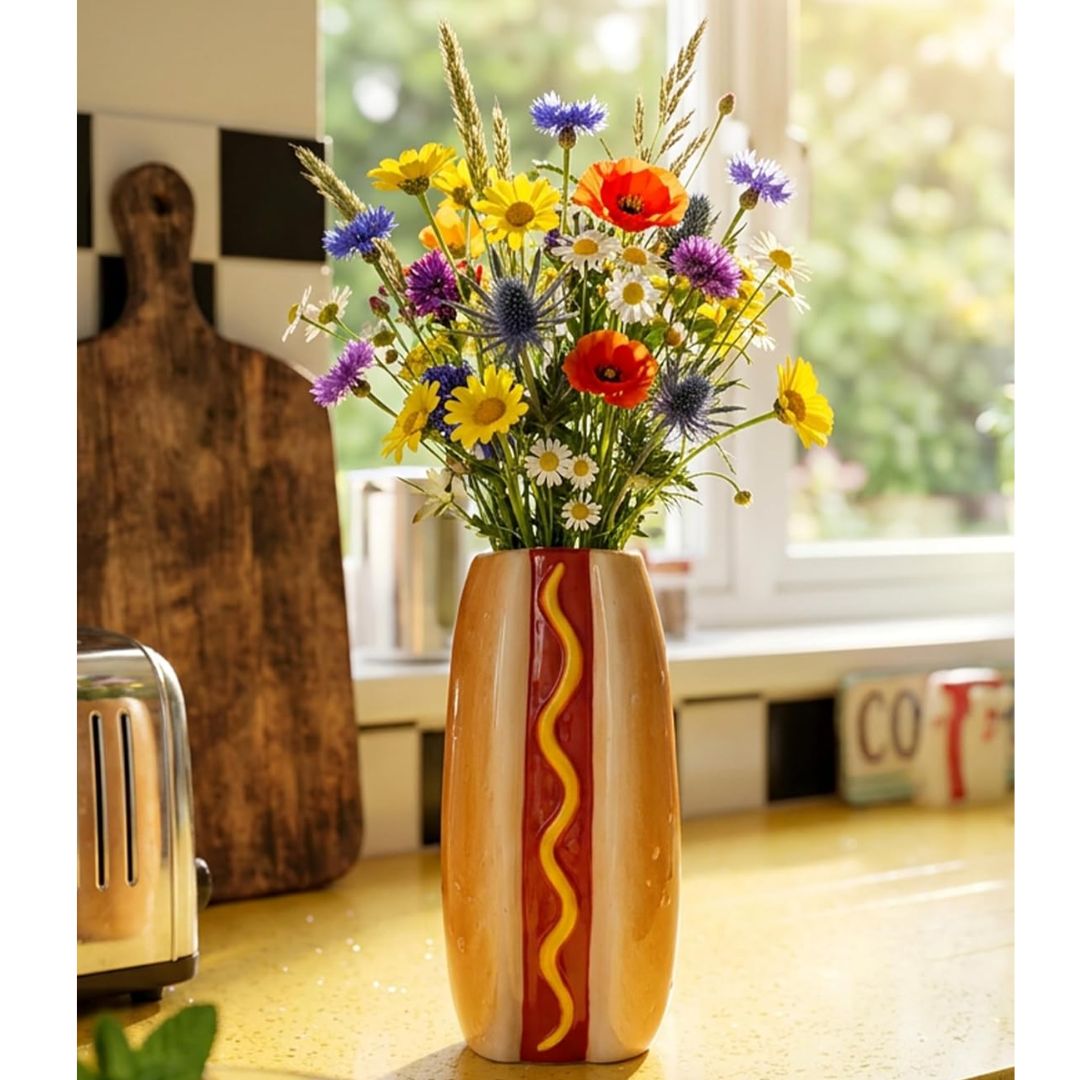A novelty ceramic vase shaped like a hot dog, filled with a colorful bouquet of wildflowers, sitting on a kitchen counter in front of a window.