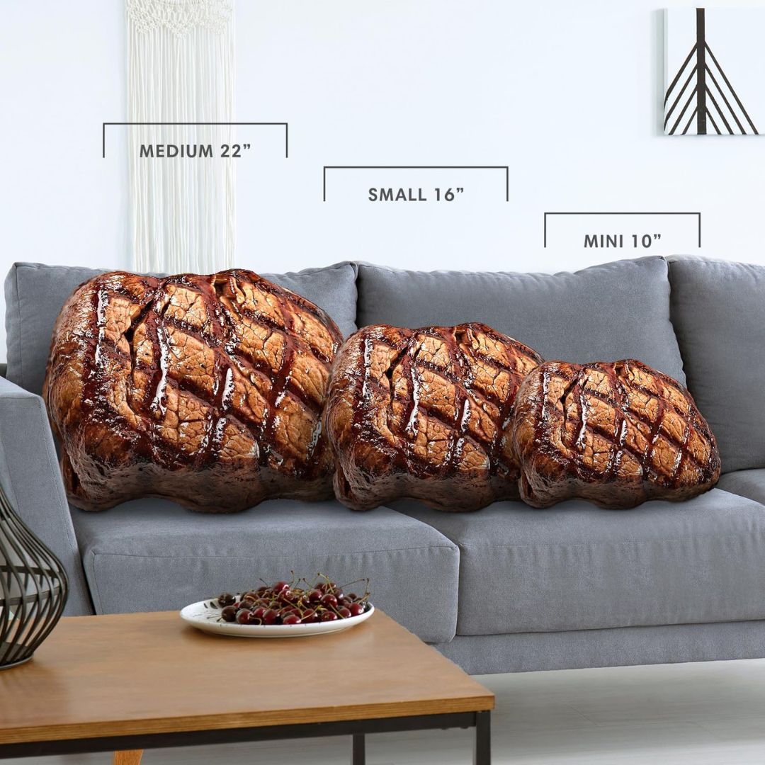 Three plush throw pillows shaped like grilled meat rests on a gray sofa in a living room setting, with size labels indicating Medium 22", Small 16", and Mini 10".