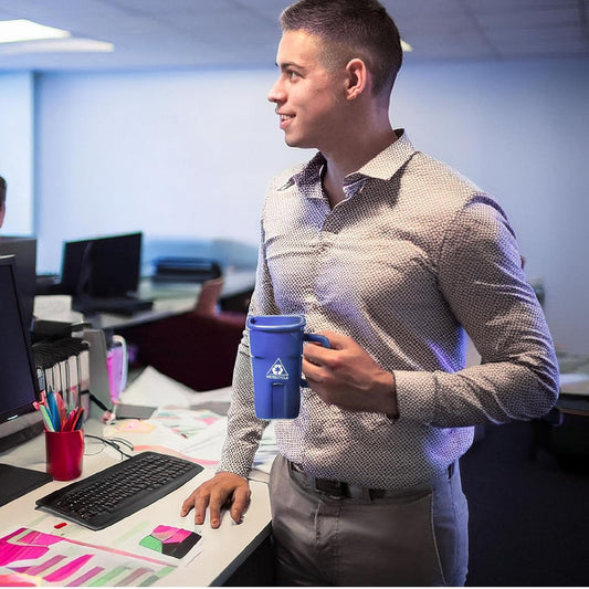 A man in an office holds a blue coffee mug designed to resemble a recycling bin. He stands by a desk with colorful documents, a keyboard, and office supplies.