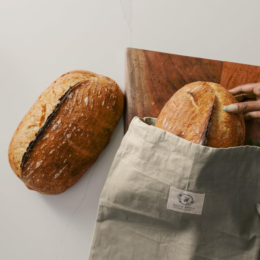 A reusable beeswax sourdough storage bag with a load of bread inside the bag with another loaf of bread sitting alongside the bagged loaf on a wooden board