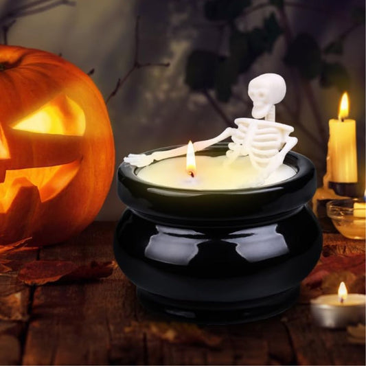 A black cauldron shaped candle with a white skeleton figurine sitting on the rim, lit with a candle, set on a wooden surface with a jack-o'-lantern and other Halloween decorations in the background.