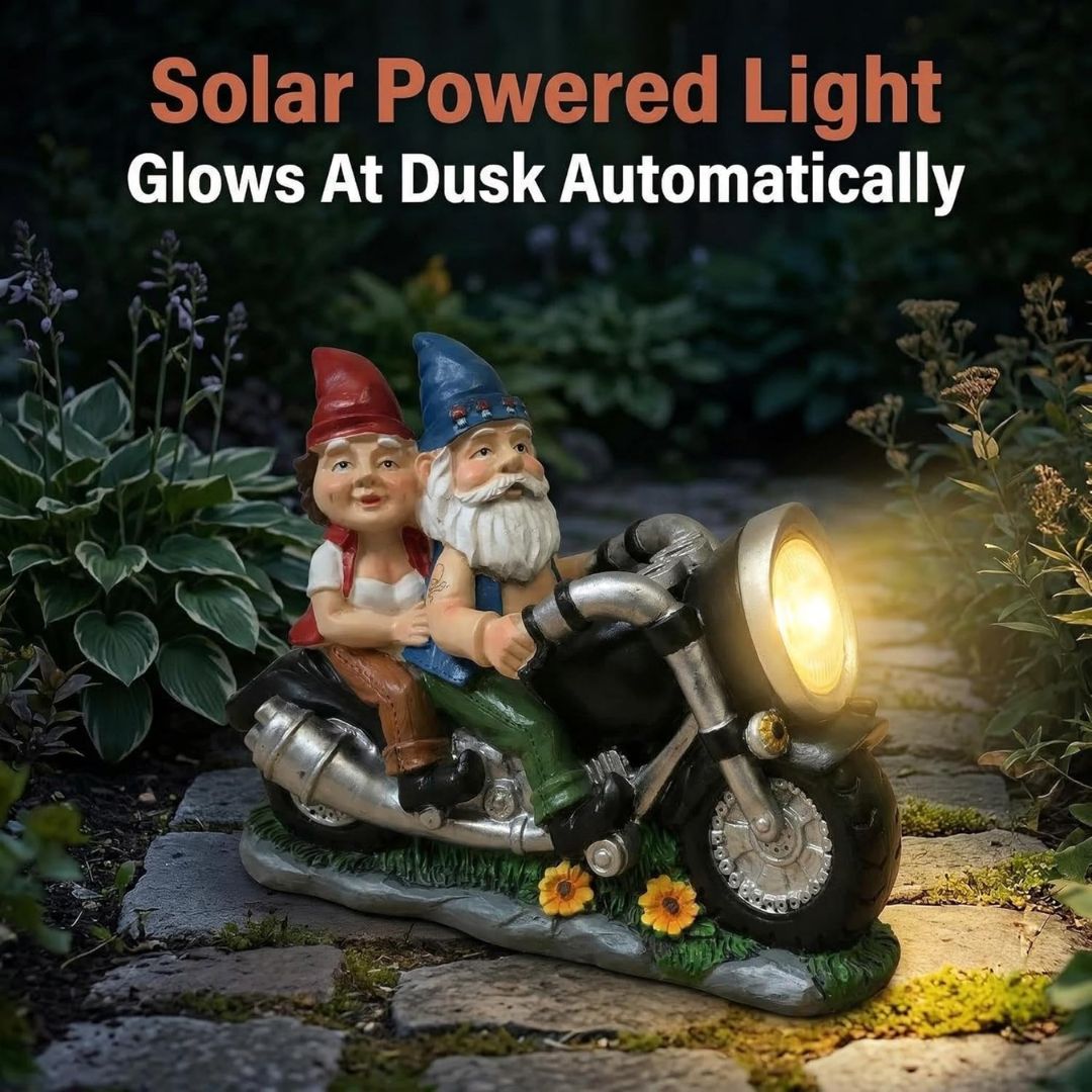 Solar powered garden light shaped like a motorbike with two garden gnomes riding it, shown glowing at dusk in a garden setting. Text overlay at the top reads 'Solar Powered Light Glows At Dusk Automatically'.