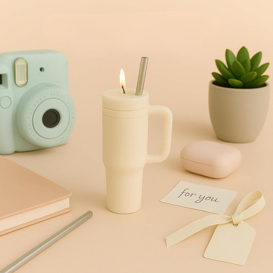 Tumbler shaped candle with a camera, notebook, and plant on a beige background