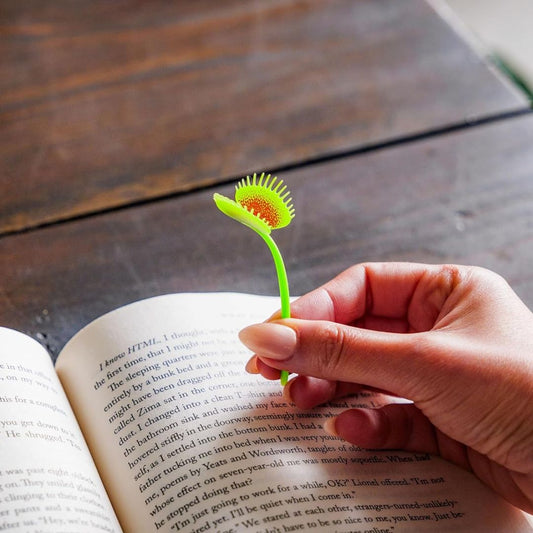 Person holding a green Venus flytrap bookmark over an open book on a wooden surface