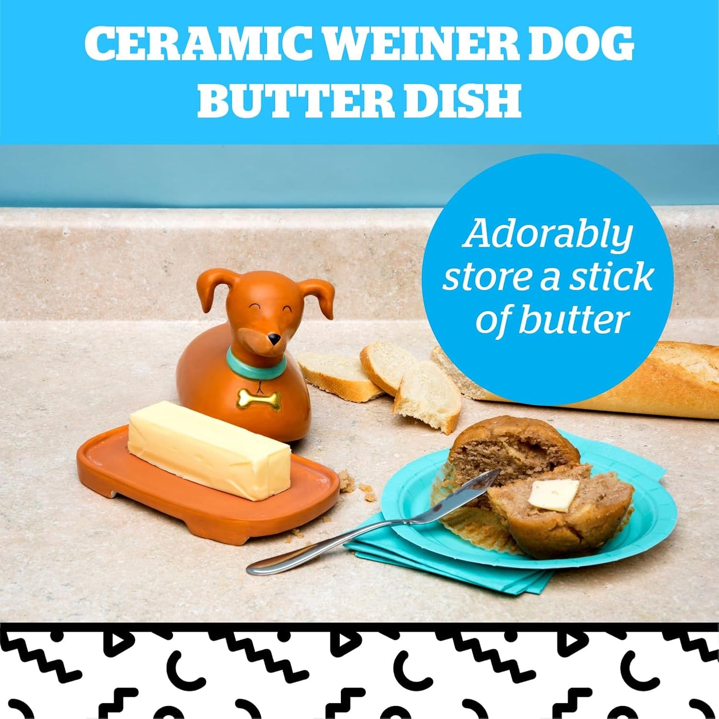 A ceramic dachshund-shaped butter dish holding a stick of butter on a brown tray, with bread, a muffin, and a plate with a buttered muffin nearby.