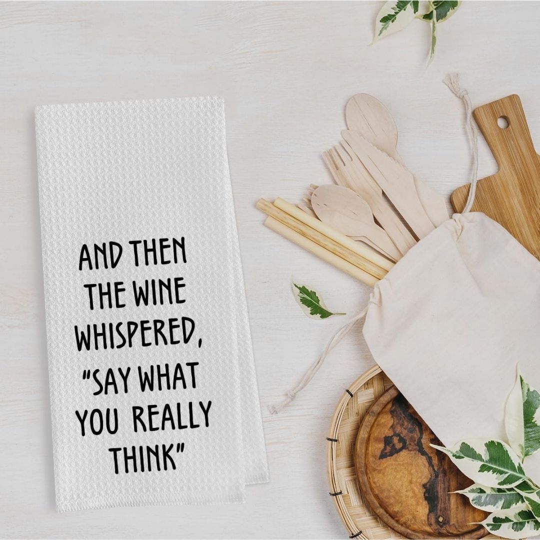 White towel with humorous text next to wooden cutlery and a bag on a light surface.