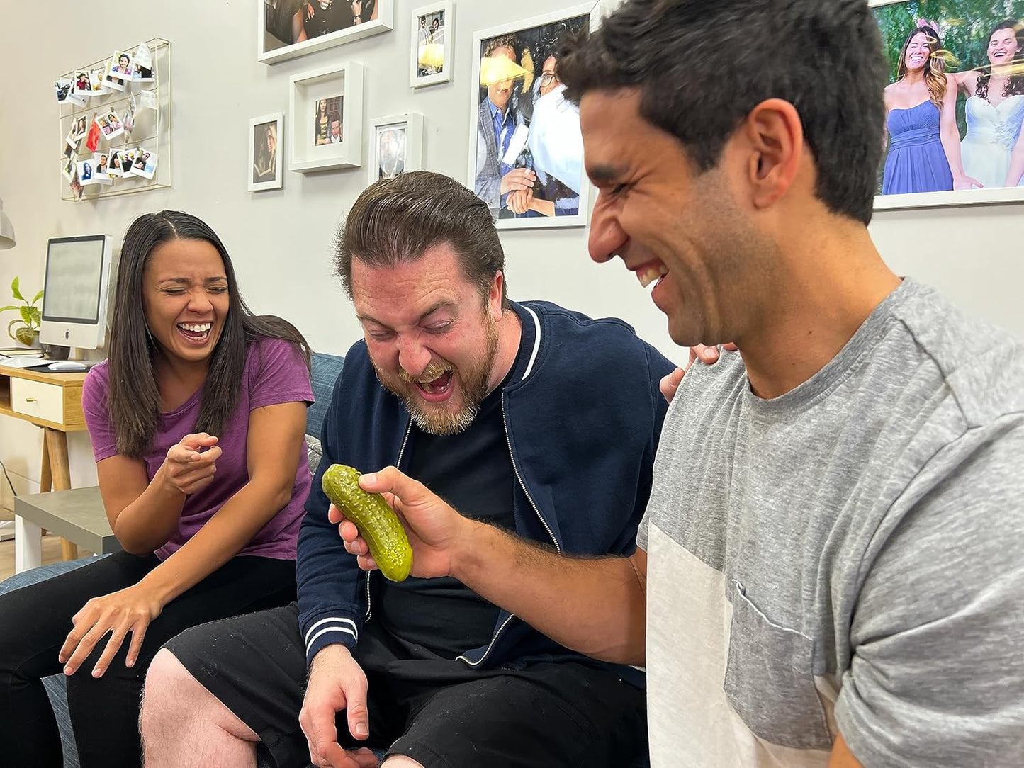 Three friends laughing together as one of them holds a yodeling pickle toy in a cozy living room setting.
