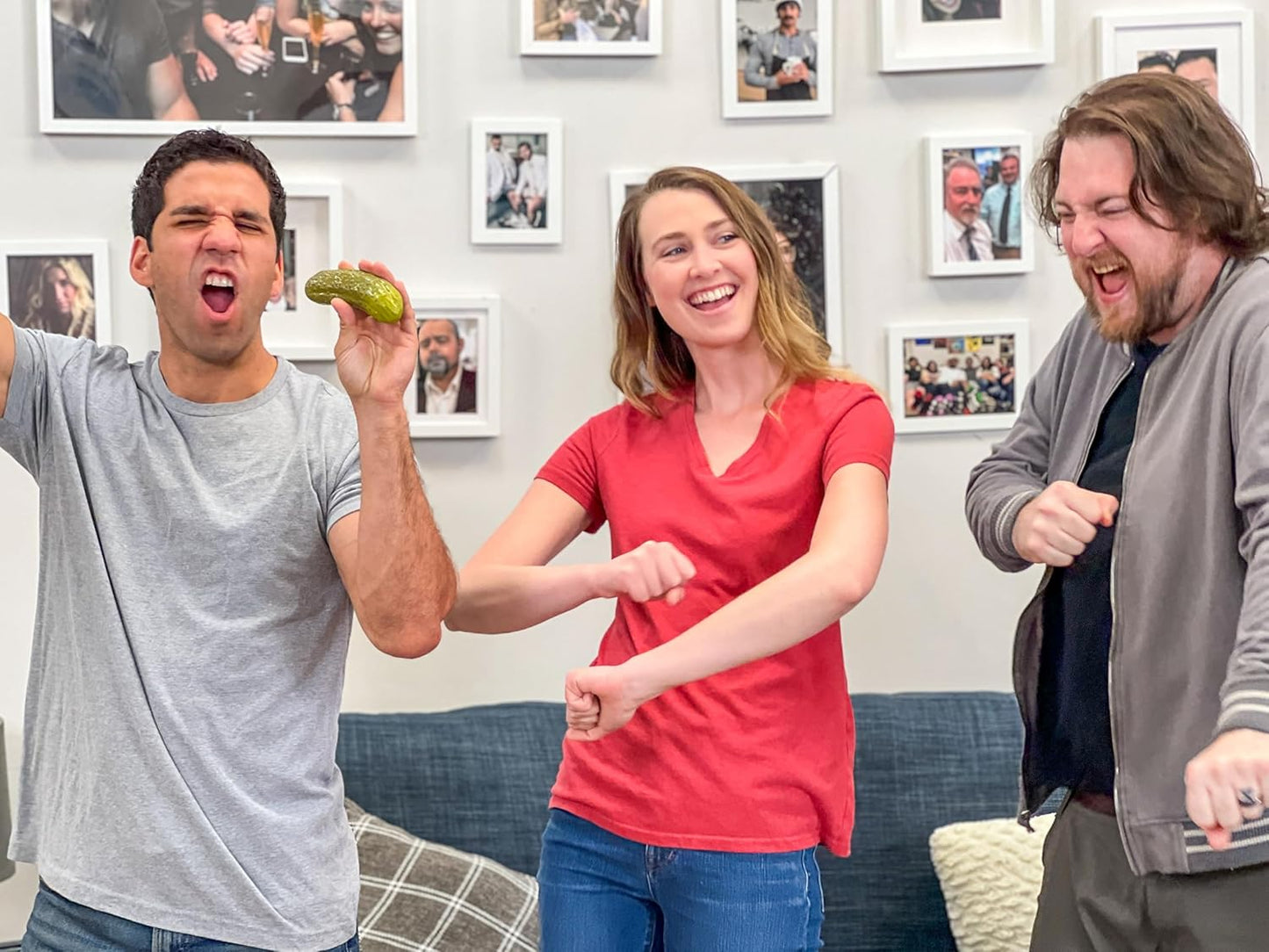 Three people dancing and laughing as one man holds up a yodeling pickle toy in a fun, lively room with framed photos on the wall.