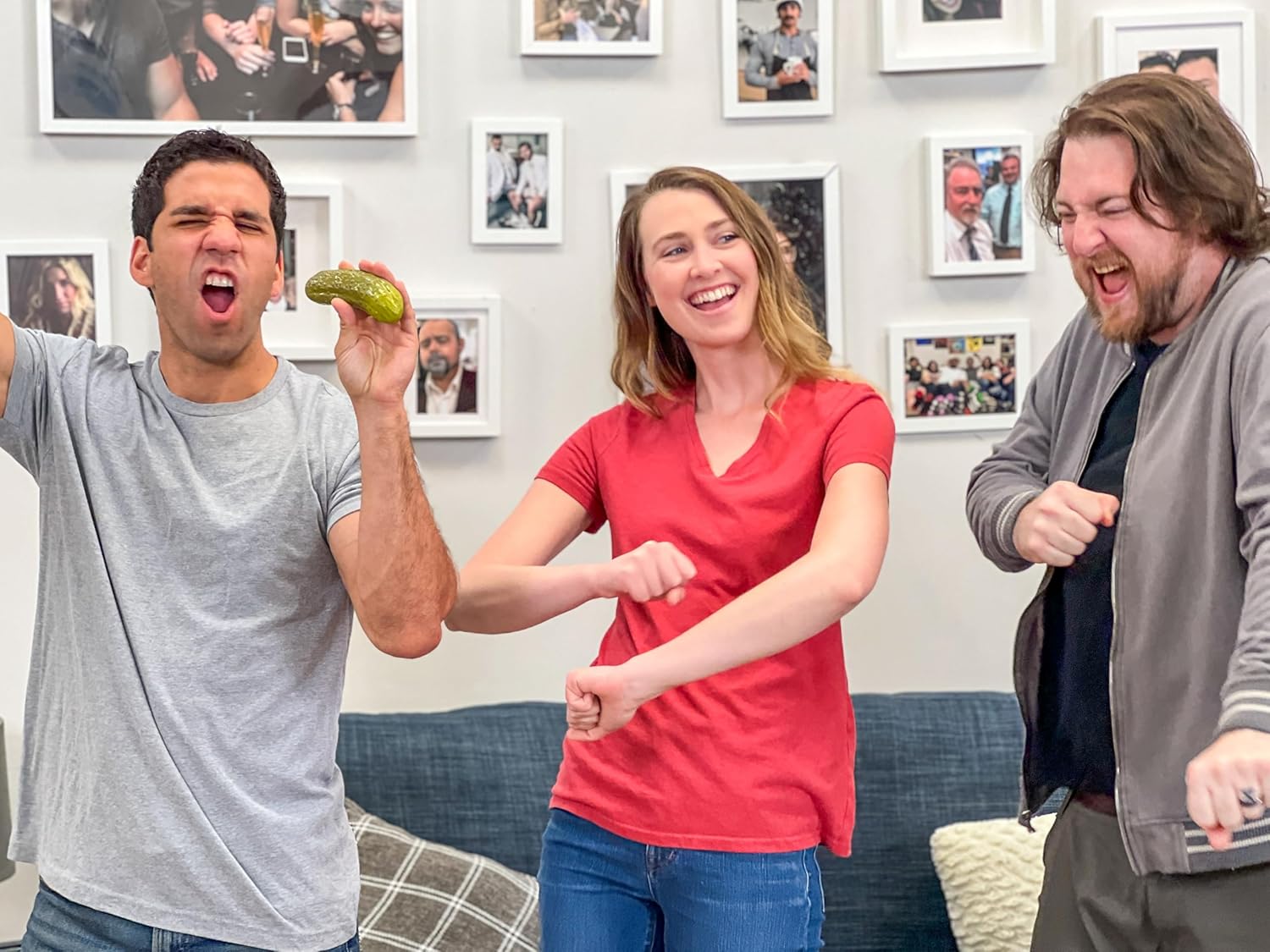 Three people dancing and laughing as one man holds up a yodeling pickle toy in a fun, lively room with framed photos on the wall.