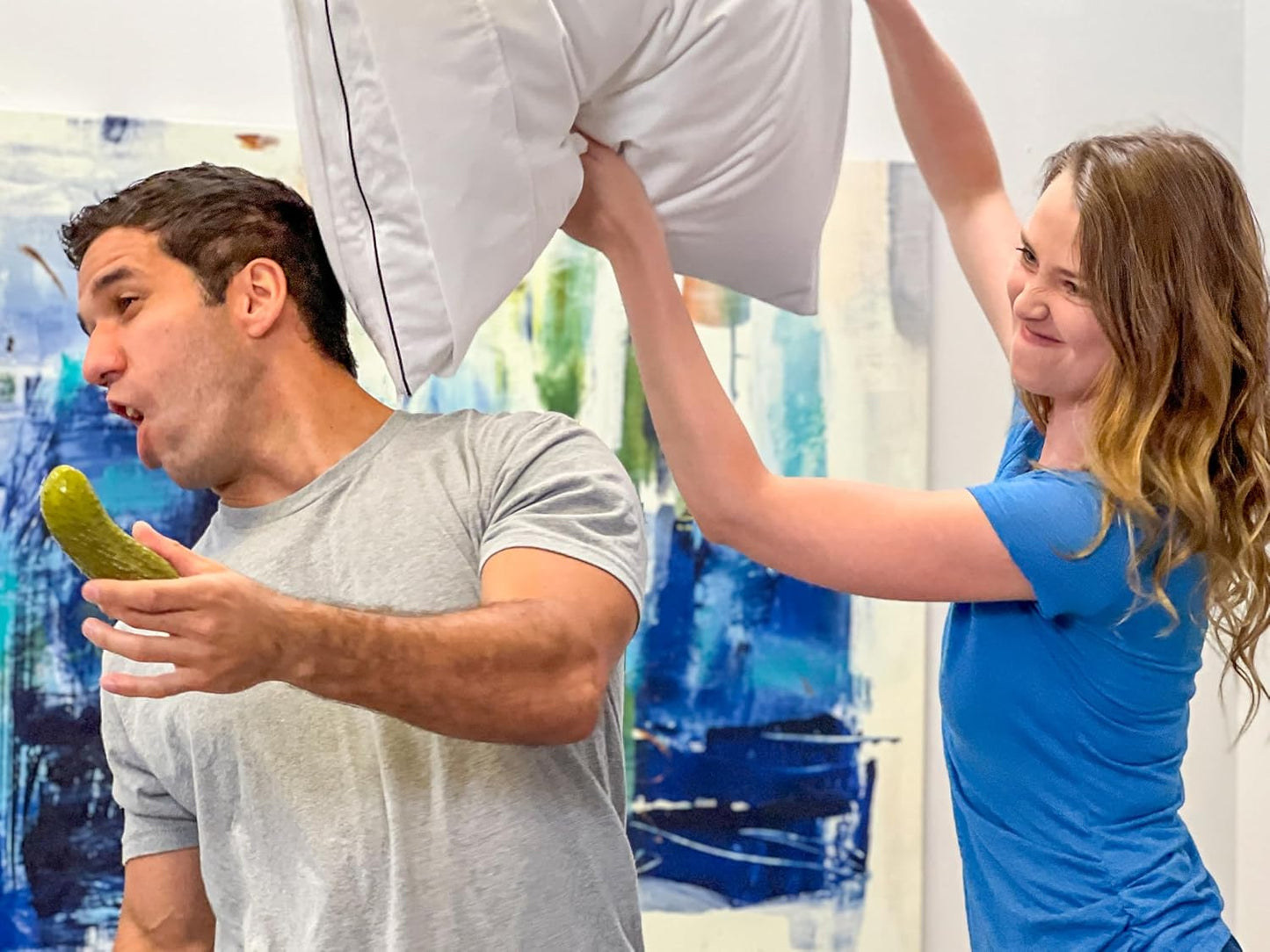 Woman playfully hitting man with a pillow while he reacts dramatically, holding a yodeling pickle toy in a brightly decorated room.