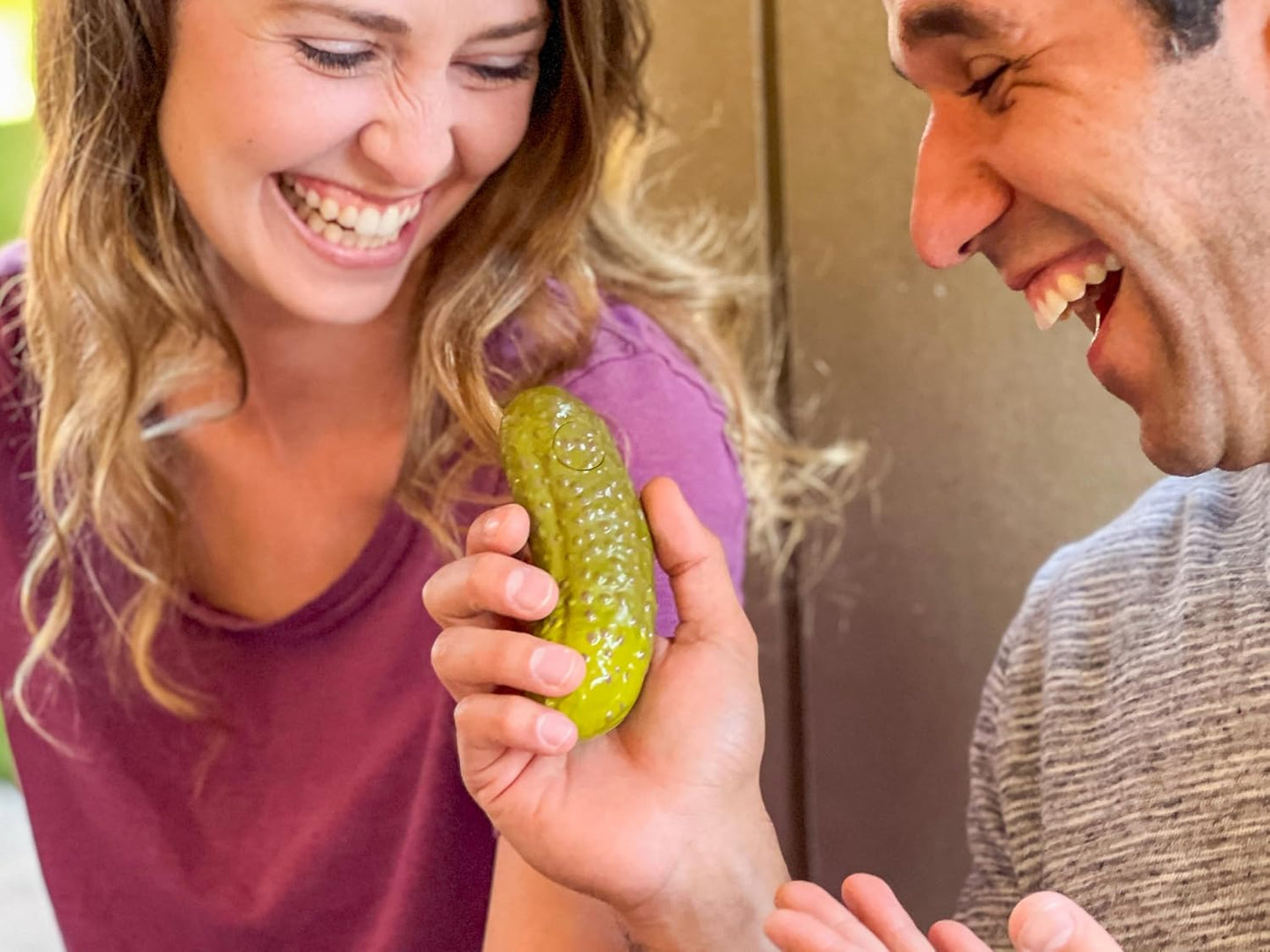 Two people laughing together while holding a green plastic yodeling pickle toy close-up.