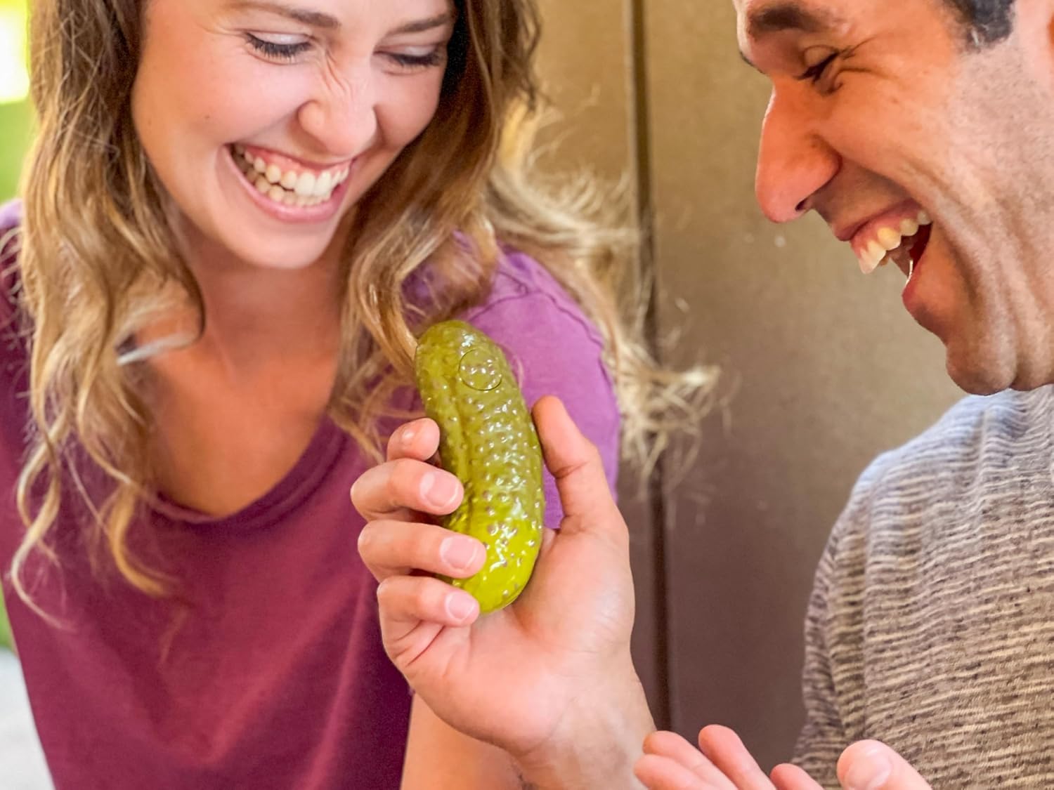 Two people laughing together while holding a green plastic yodeling pickle toy close-up.