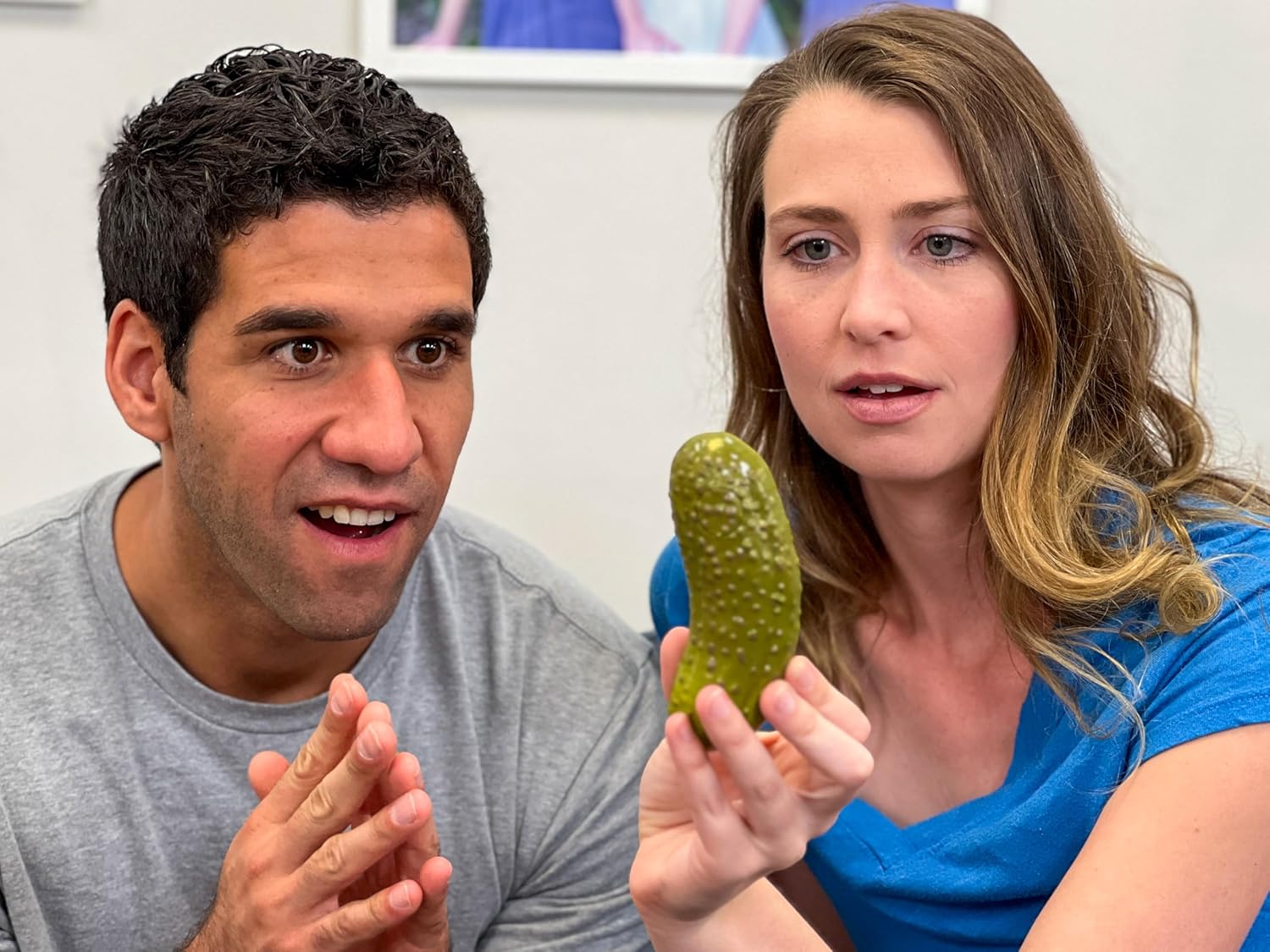 A man and woman stare intently at a green plastic yodeling pickle toy, both looking surprised and fascinated by it.