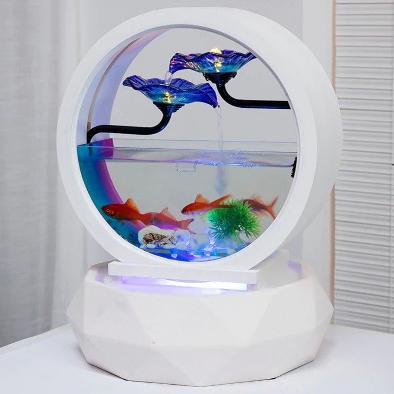 A decorative aquarium sitting on a white table. The aquarium features a waterfall with goldfish inside the tank. There are 2 blue glass flowers on top and water is cascading down the flowers and into the aquarium. The base is an air purifier. The aquarium and base are both white and circular. The air purifier is on and clean air can be seen coming out of it.